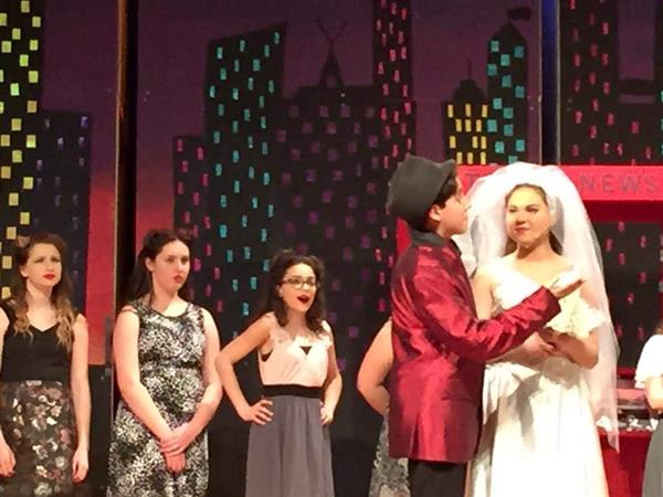 A young boy in a red suit and black hat embraces a girl in a white dress on stage, surrounded by other performers. The backdrop features cityscapes.