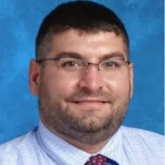 A man in a light blue shirt with a plaid tie poses against a textured gray background, exuding a professional and approachable demeanor.