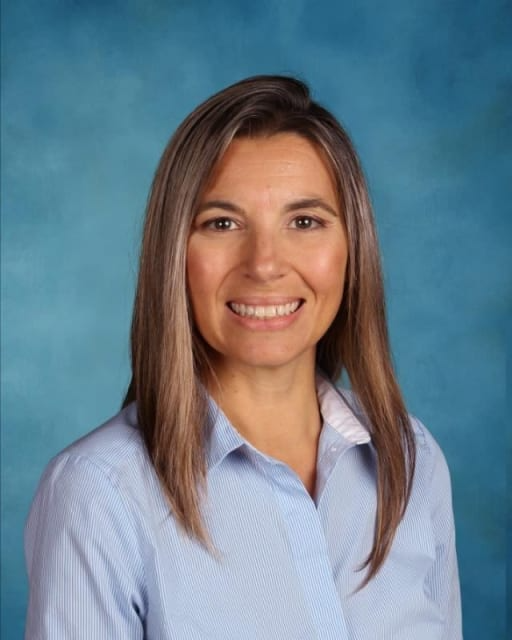 A woman wearing a blue shirt with a white collar, standing confidently and smiling at the camera.