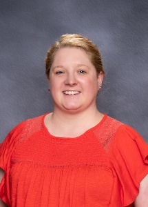 A woman with short blonde hair is smiling, wearing a bright red blouse, against a gray background.