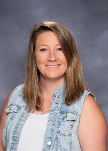 A smiling woman with long brown hair wearing a denim vest over a white shirt, set against a gray background.