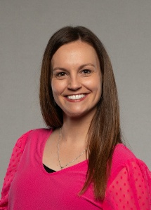 A woman with long brown hair smiles while wearing a bright pink top with textured sleeves against a neutral gray background.
