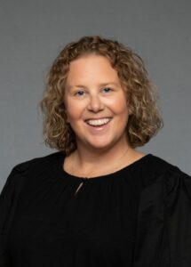 A smiling woman with curly hair wearing a black top, set against a gray background.