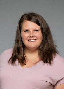 A woman with long brown hair smiles at the camera, wearing a light pink shirt against a gray background.