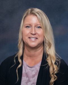 A woman with long, wavy blonde hair smiles at the camera, wearing a black cardigan over a light purple top, set against a blurred dark background.
