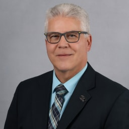 A middle-aged man with gray hair and glasses, wearing a black suit and a light blue shirt with a patterned tie, poses against a neutral gray background.