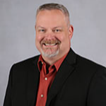 A smiling man with short gray hair and a beard, wearing a black blazer over a red shirt, poses against a light gray background.