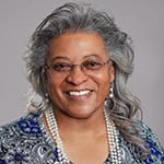 A smiling woman with gray hair styled in loose waves, wearing glasses and a patterned blue dress, adorned with multiple strands of pearls.