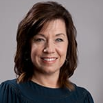 A woman with shoulder-length brown hair, wearing a dark blue blouse, smiles at the camera against a gray background.