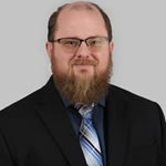 A man with a beard wearing glasses, a black suit, and a blue and white striped tie, standing against a gray background.