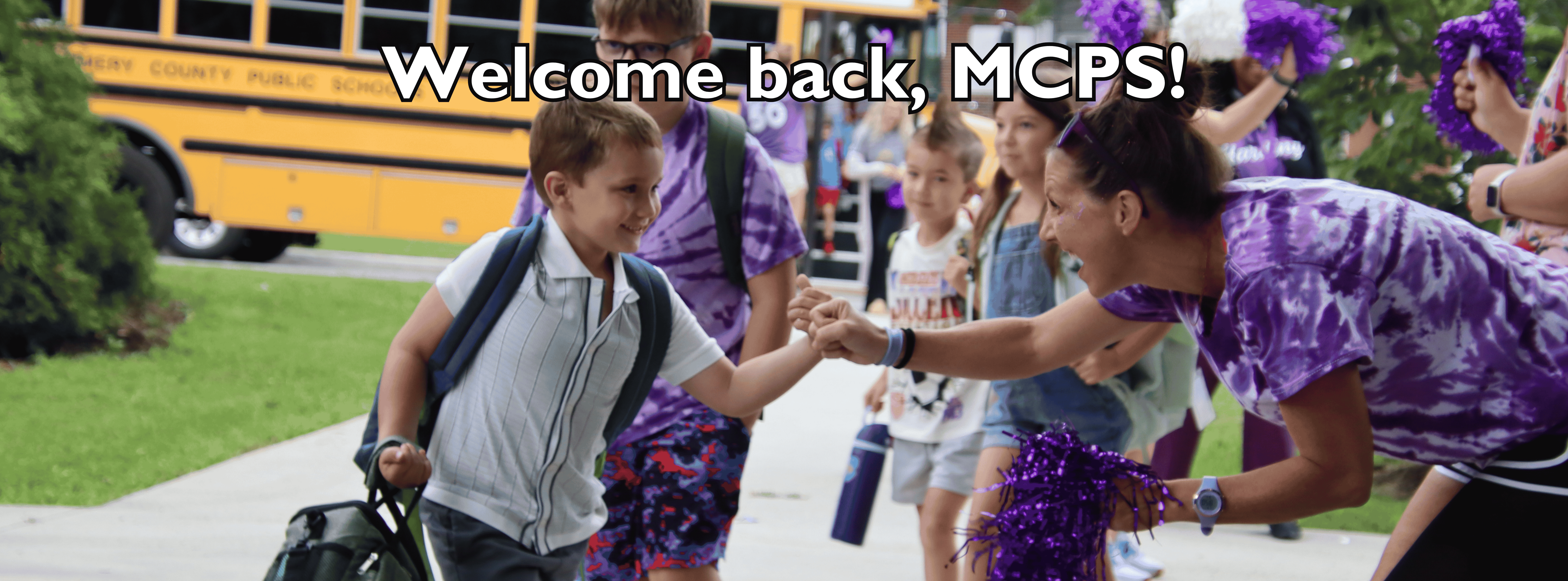 A young boy with a backpack and lunch bag fist-bumping a cheerful woman in a purple tie-dye shirt as he arrives at school. In the background, other students step off a yellow school bus that reads "Montgomery County Public Schools," welcomed by people holding purple pom-poms. The text overlay centered near the top reads, "Welcome back, MCPS!"