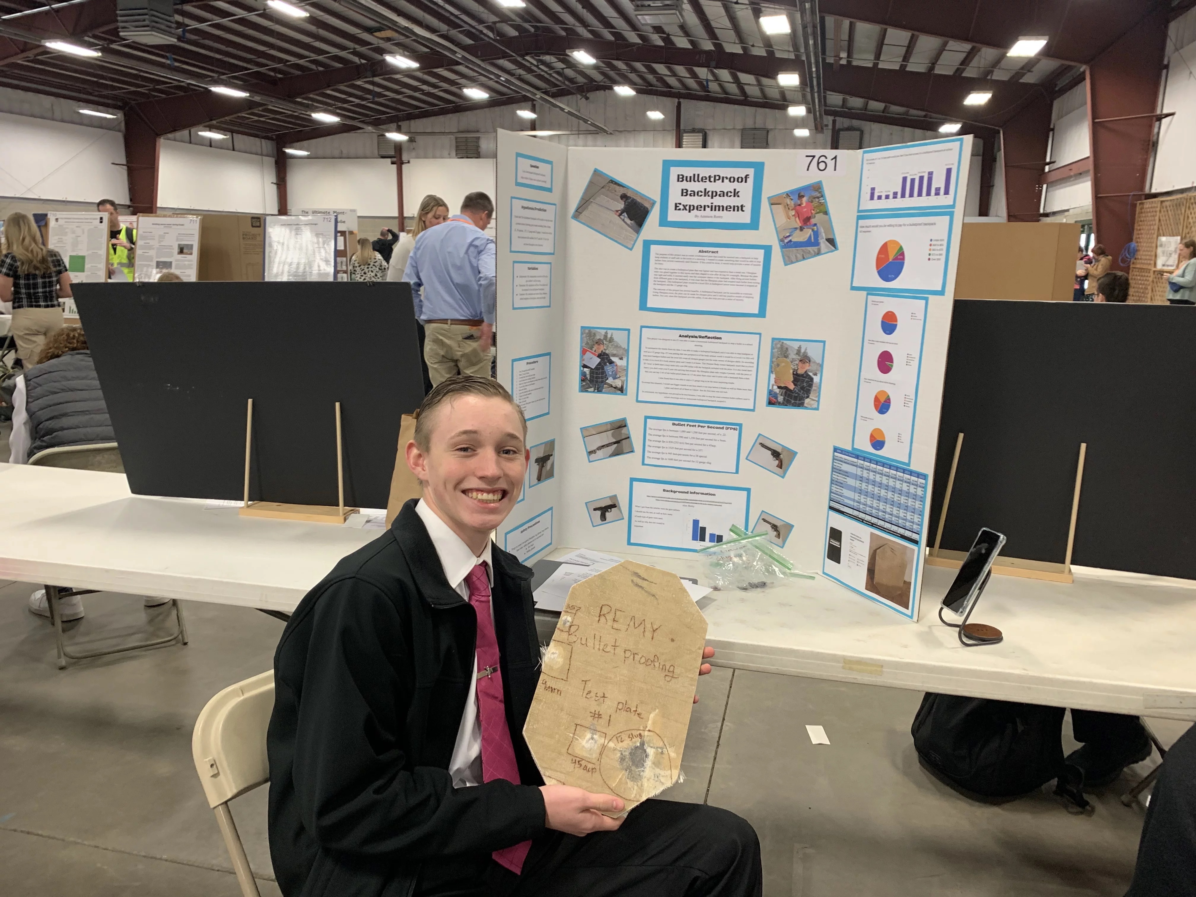 A young man proudly presents a plaque at an event, standing in front of a table with information displayed on it.