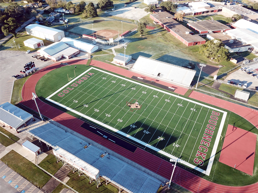 An aerial view of a football field surrounded by school buildings and green spaces, featuring a well-marked turf field and bleachers.