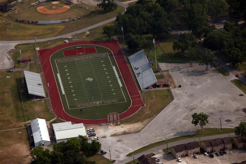 An aerial view of a sports field with a football pitch, surrounded by bleachers and a parking area, set in a grassy landscape.