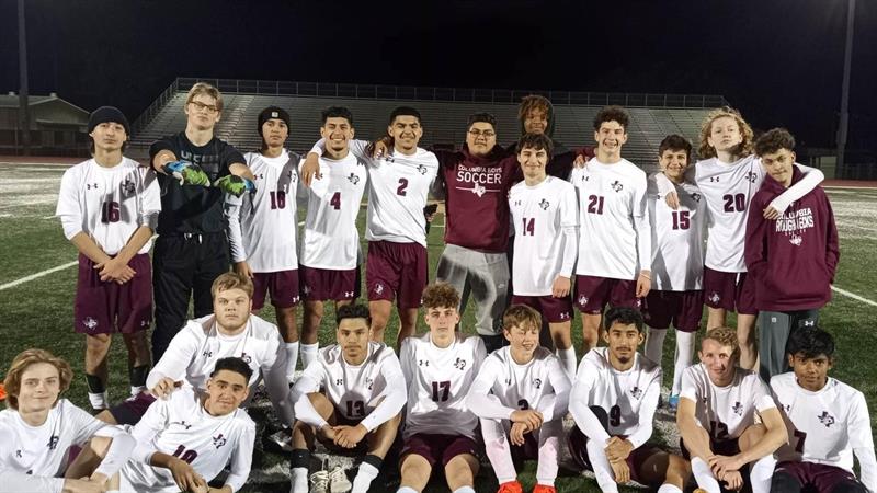 A group photo of a youth soccer team, wearing white and maroon uniforms, posing together on a field at night, with some players kneeling and others standing behind them, all smiling and celebrating.