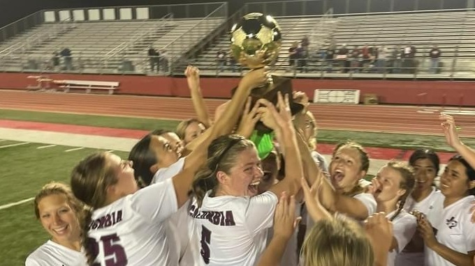 A group of female soccer players joyfully lift a trophy above their heads in celebration on a sports field, with empty bleachers in the background.