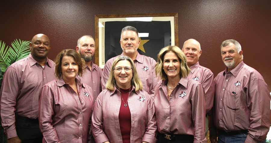 A group of eight individuals, dressed in matching maroon shirts, pose together in an office setting with a framed star emblem in the background.