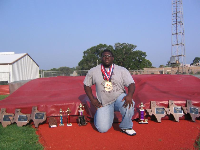 A man kneels on a track field next to several trophies, wearing medals around his neck, with a high jump mat in the background.