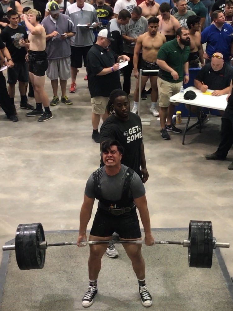 A weightlifter is performing a deadlift with a heavy barbell while being observed by a crowd of spectators and officials in a gym setting.