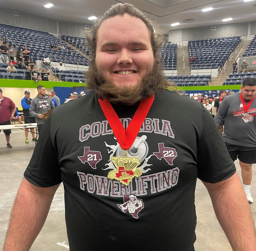 A smiling young man with a beard wearing a black t-shirt that says 'Columbia Powerlifting' and a medal around his neck, standing in a gymnasium filled with spectators.