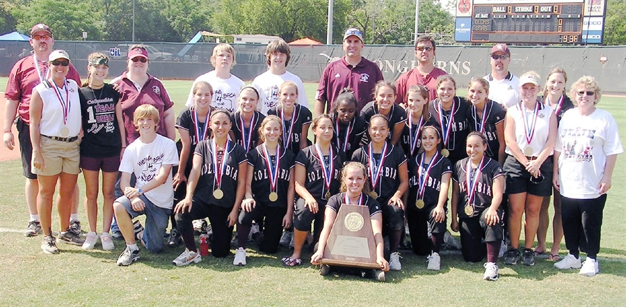 A group photo of a softball team and their coaches celebrating with medals and a trophy on a field.