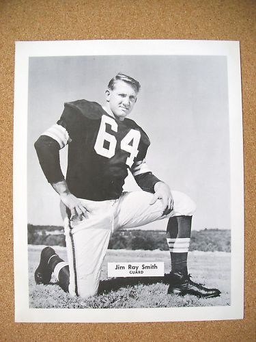 A black and white photograph of a football player kneeling on one knee, wearing a dark jersey with the number 64 and white pants, with a field in the background.