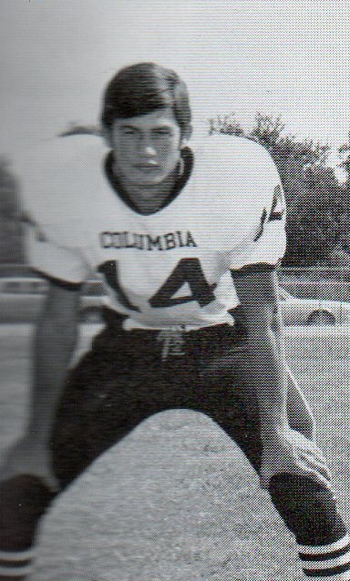 A young male football player in a Columbia jersey, number 14, is posed in a crouched stance on a football field, with trees and cars visible in the background.