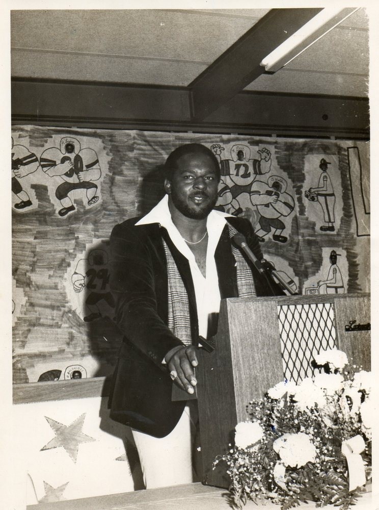 A man stands at a podium with a microphone, wearing a black jacket and white shirt, in front of a colorful mural depicting various figures and flowers on the table.