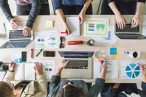 An overhead view of a collaborative workspace with multiple people using laptops, surrounded by documents, charts, and office supplies on a table.