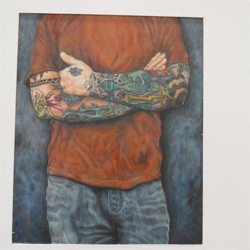 A person with colorful tattoos on their arms is standing with their arms crossed, wearing an orange shirt and blue jeans.