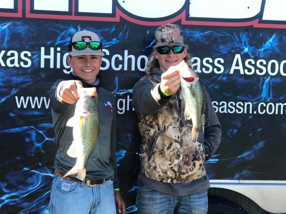 Two young anglers proudly display their catches of bass while standing in front of a trailer with a fishing association logo.