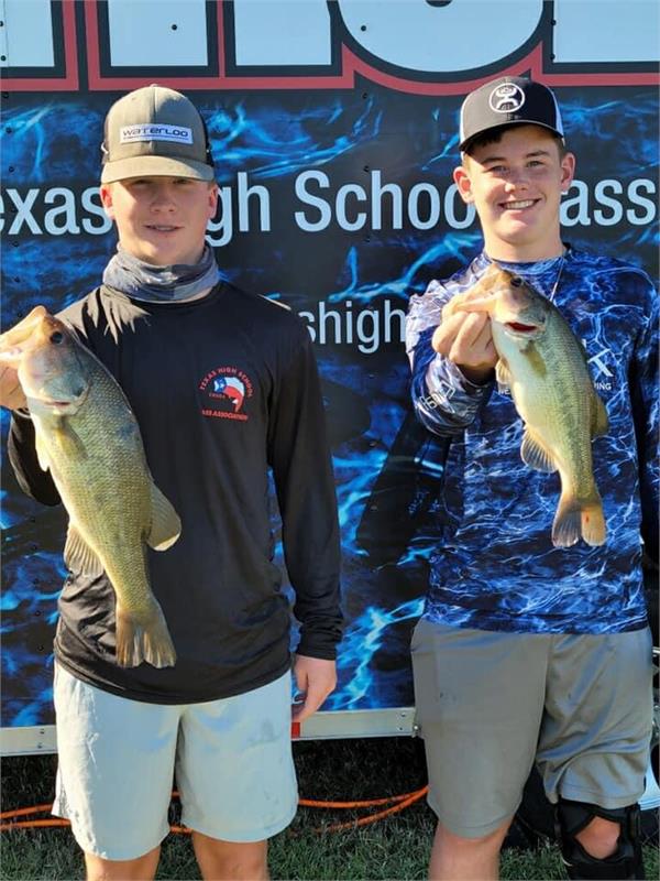 Two young anglers proudly display their catches of large bass while standing in front of a promotional banner for a Texas high school fishing event.