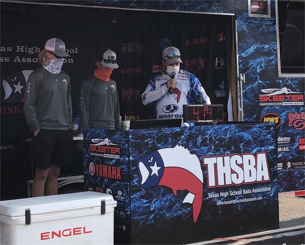 Three individuals, two wearing gray shirts and caps, stand beside a podium with a banner displaying the Texas High School Bass Association (THSBA) logo, while one person speaks into a microphone.