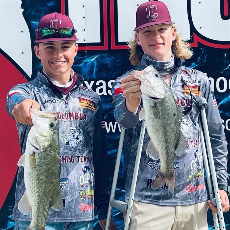 Two young anglers proudly display their catches of large bass while standing in front of a promotional backdrop.