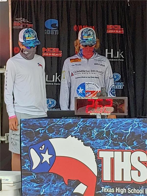 Two young anglers wearing masks and caps stand beside a podium displaying a fish-themed backdrop at a Texas High School fishing event.