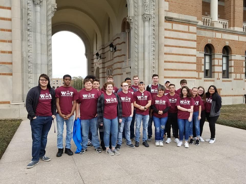 A group of students wearing matching maroon shirts stands in front of a large archway of a historic building, posing for a photo.