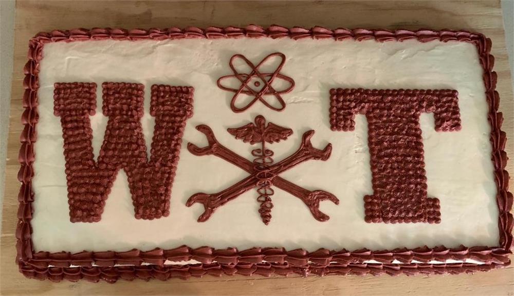 A rectangular cake decorated with the letters 'W', 'T', and a central emblem featuring a wrench and caduceus, all outlined in red icing on a white frosting background.