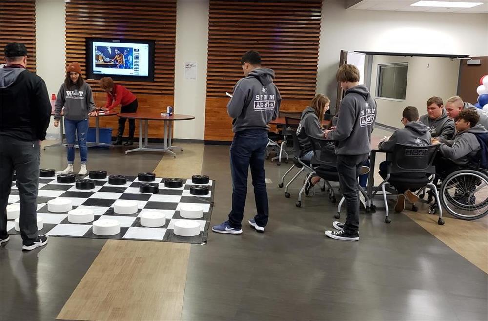 A group of people is gathered around a large checkers board set up on the floor, with some participants standing and others seated, including individuals in wheelchairs, while a presentation is visible on a screen in the background.