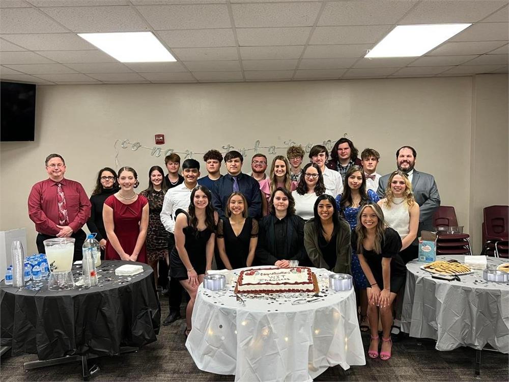 A group of students and adults gathered together for a celebration, posing in front of a decorated table with a cake and refreshments in a well-lit room.