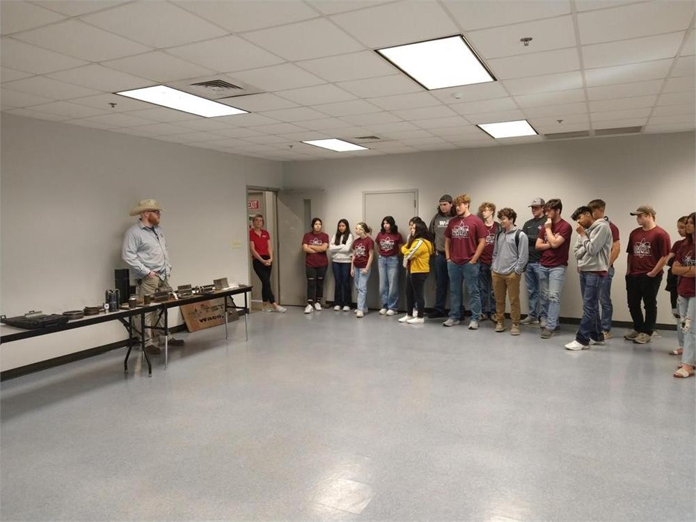 A group of students in matching shirts attentively listens to a speaker standing beside a table with various items in a spacious, well-lit room.