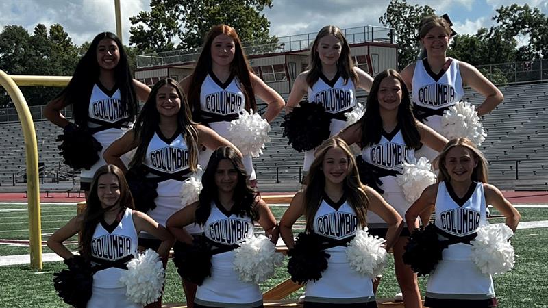 A group of cheerleaders poses together on a football field, wearing matching uniforms and holding pom-poms, with a stadium in the background.