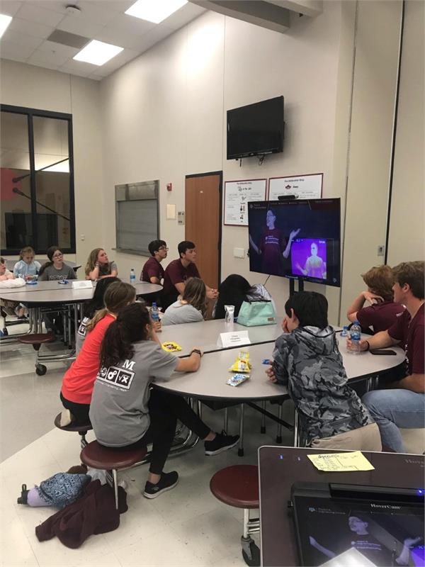 A group of students seated at tables in a classroom, attentively watching a presentation on a television screen.