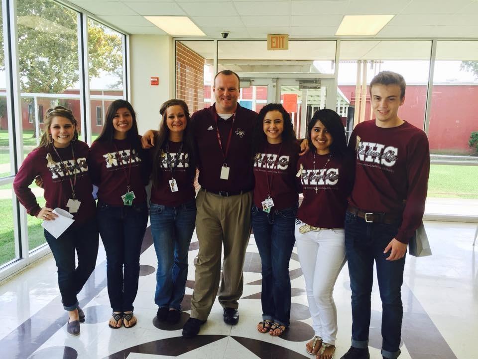 A group of seven individuals, wearing matching maroon shirts with the text 'WHC' on them, stand together in a brightly lit hallway with large windows in the background.