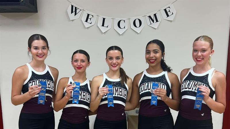 Five cheerleaders stand together, smiling and holding blue ribbons, in front of a 'WELCOME' banner.