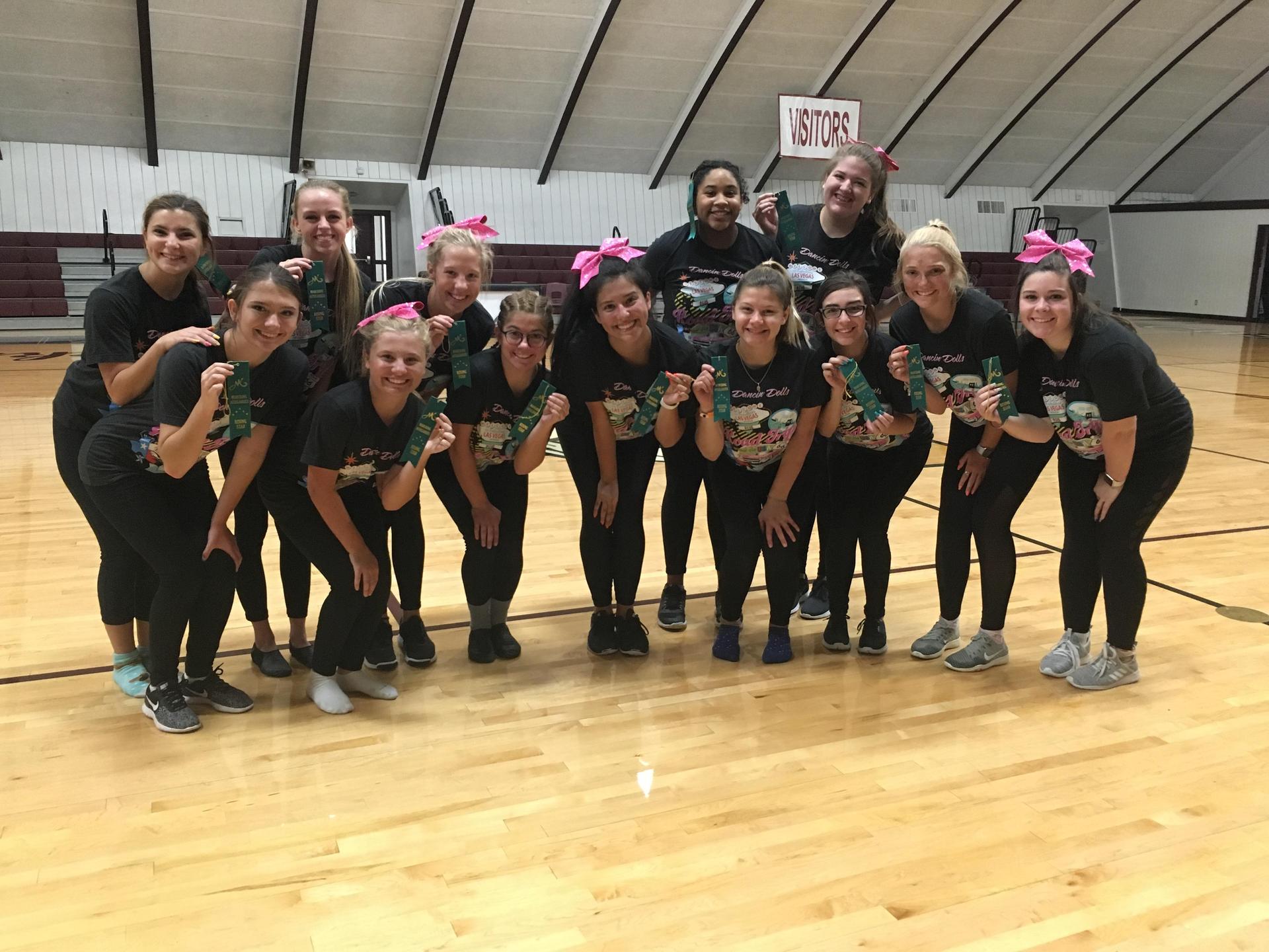 A group of cheerleaders in matching black shirts and pink bows pose together on a gymnasium floor, smiling and showing their cheerleading spirit.