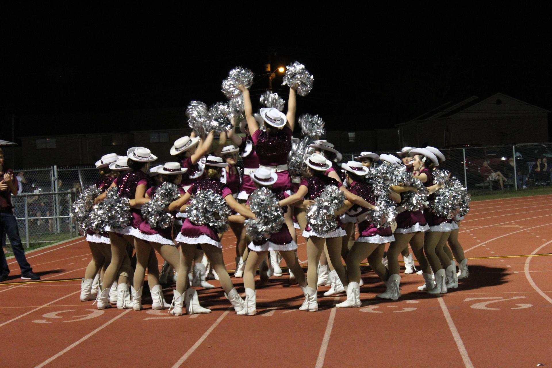 A group of cheerleaders in matching outfits perform a pyramid formation on a track field at night, with one cheerleader elevated at the top holding pom-poms.