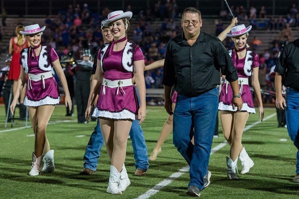 A group of performers in pink and white costumes walks on a football field, accompanied by a man in a black shirt, during a nighttime event.