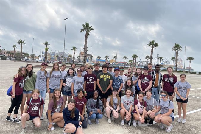 A large group of students stands together in a parking lot, with palm trees and a cloudy sky in the background.