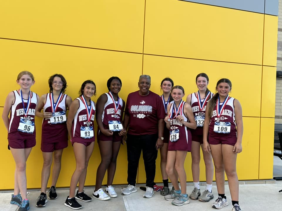 A group of eight female runners wearing athletic uniforms and medals stand together with a coach in front of a bright yellow wall, celebrating their achievement.