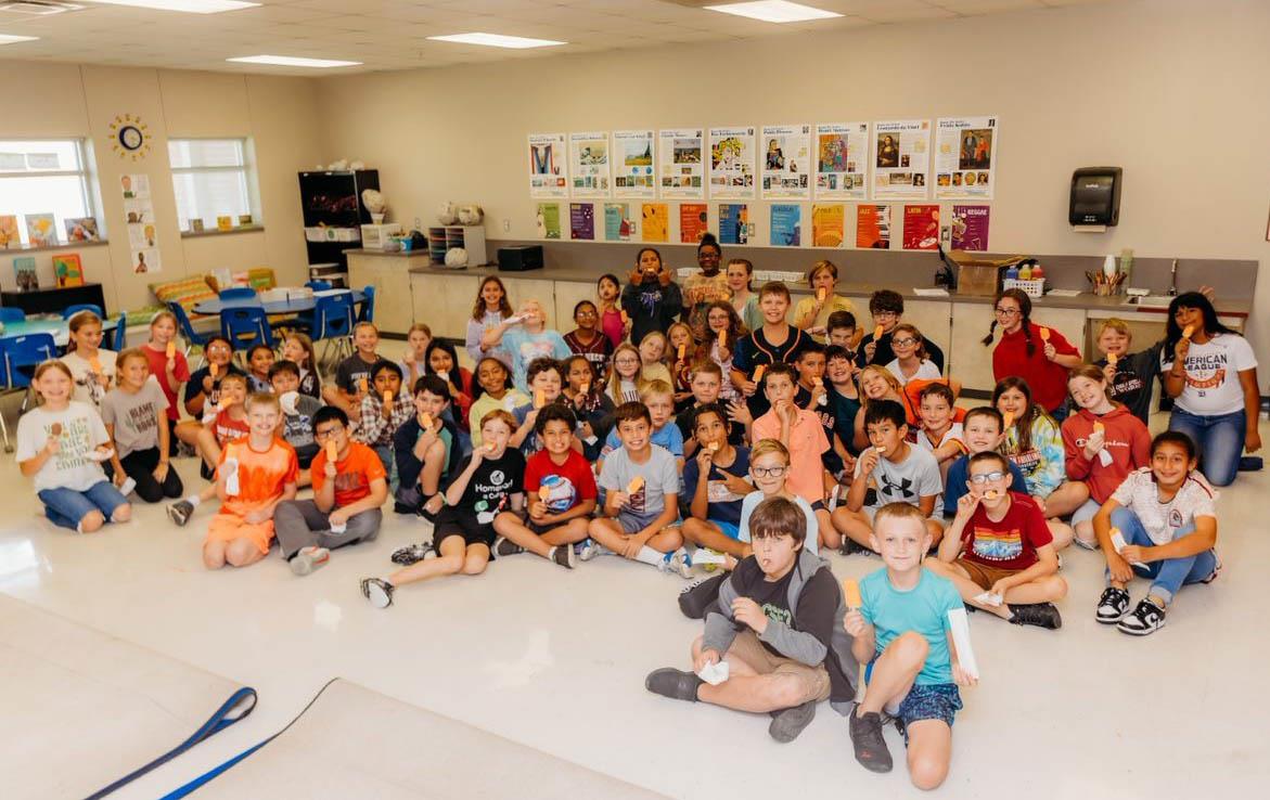 A large group of children and adults are gathered together in a classroom, smiling and posing for the camera, with colorful posters on the wall behind them.